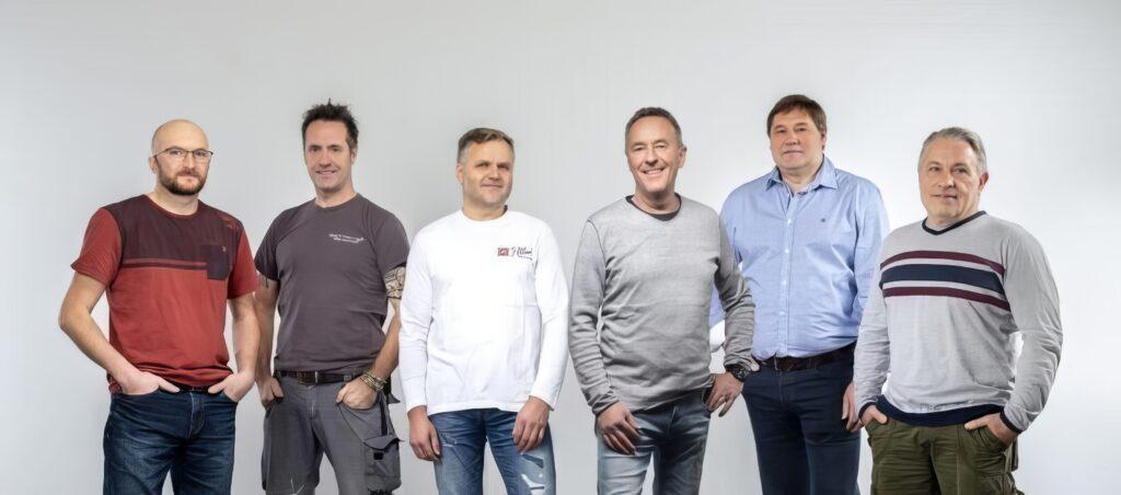 Sechs Personen lächeln vor einem neutralen Hintergrund.